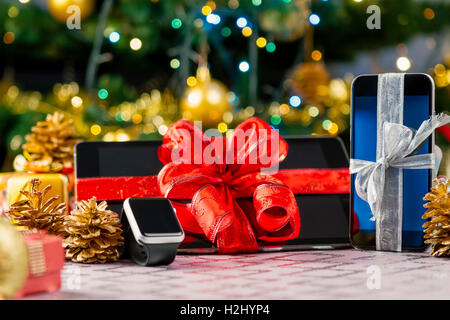 Tablet PC, smartphone e smartwatch con doni e decorazioni nella parte anteriore dell'albero di Natale. Focus sullo smartphone. Foto Stock