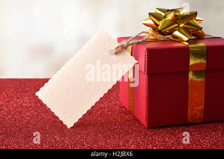 Confezione regalo rossa con golden nastro con etichetta vuota appeso sul colore rosso brillante tavolo e sfondo bianco. Vista frontale Foto Stock