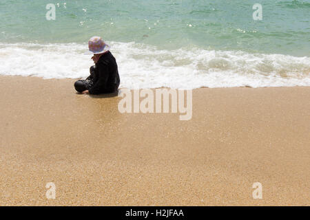 Thai donna seduta sulla spiaggia in un abito nero e un grande cappello, picking cozze in terra-wash. Spazio per testi Foto Stock