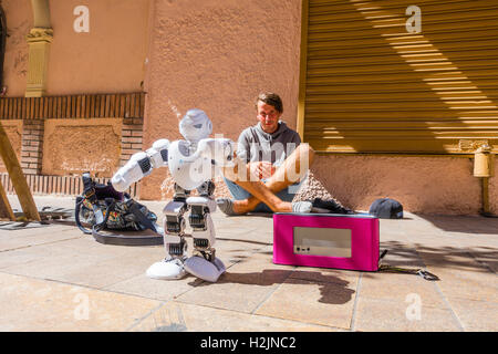 Una ventina di qualcosa di maschio si siede sul marciapiede con il suo robot di eseguire per passanti come egli tenta di panhandle per denaro. Foto Stock