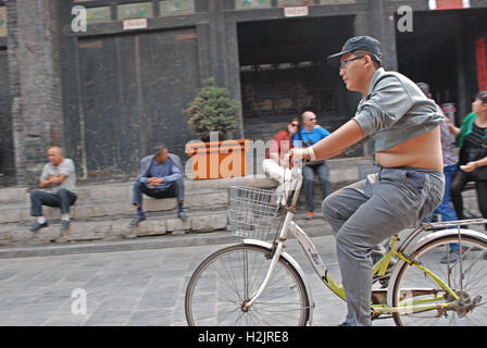 Un locale uomo cinese cicli attraverso la strada principale di Pingyao, la Cina con il suo ventre esposta. Foto Stock