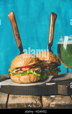 In casa hamburger di manzo con pancetta croccante, anelli di cipolla, sottaceti, verdure fresche e vetro sul grano portano a servire la tavola di legno Foto Stock