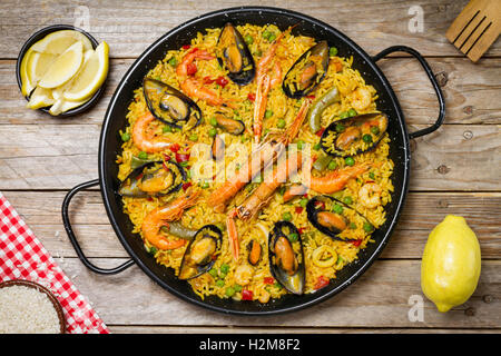 La paella spagnola su una tavola di legno con limone fresco e un bianco e rosso tovaglia e una forcella di legno Foto Stock