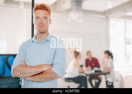 Ritratto di fiducioso giovane imprenditore in piedi in ufficio con le braccia incrociate e colleghi riuniti in background. Foto Stock