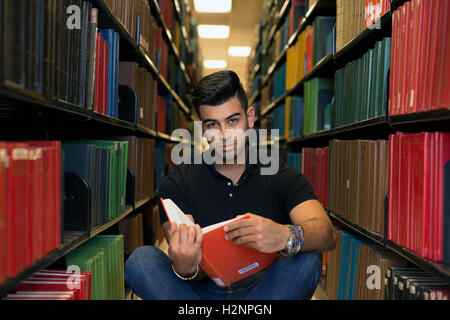 Collegio maschile studente visto nella libreria, seduta sul pavimento tra le righe di libri. Egli è in possesso di un libro, guardando la fotocamera. Foto Stock