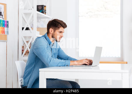 Vista laterale di un pensieroso bello barbuto imprenditore casual seduto con il portatile in ufficio Foto Stock