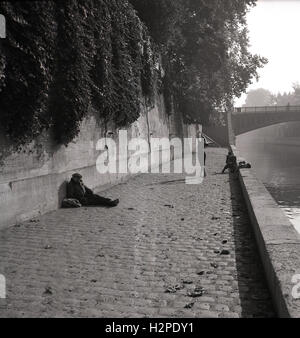 Degli anni Cinquanta, una moda sparare sulle rive del Fiume Senna, Parigi, Francia, fotografo di scattare le foto di una femmina in tacchi e condire con un vagabondo seduto su il ciottolo contro una parete. Foto Stock