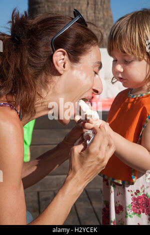 Donna di mangiare godendo di ghiaccioli bianco gelato da tre anni bambino biondo mani, con orange bib per esterno Foto Stock