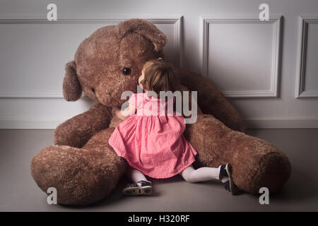 Orizzontale vista posteriore della bambina in abito di ping abbracciando una molto grande orsacchiotto seduto sul pavimento Foto Stock