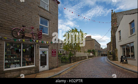 Ciottoli deserta strada costeggiata da negozi e case in sonnolento villaggio storico di Hawes, sotto il cielo blu, nello Yorkshire, Inghilterra Foto Stock