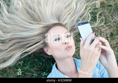 Giovane e bella bionda sdraiati sull'erba e tenendo selfie Foto Stock