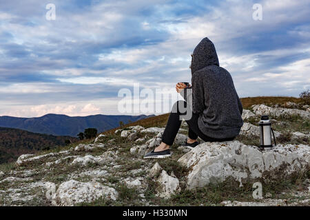 Unidentified donna che indossa hoodie seduto sulla cima della montagna tenendo un thermos la tazza di tè e godersi il tramonto Foto Stock