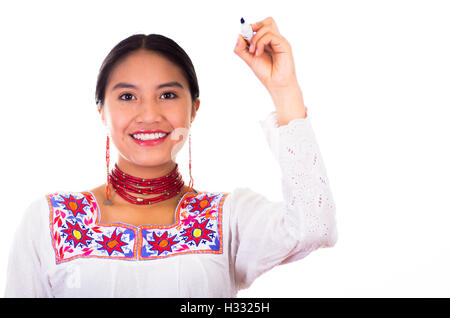 Affascinante giovane donna indossa tradizionale camicetta andina con ricami colorati, matching red collana e orecchini, di fronte alla fotocamera durante la scrittura in aria utilizzando marker, sorridendo felicemente, bianco di sfondo per studio Foto Stock