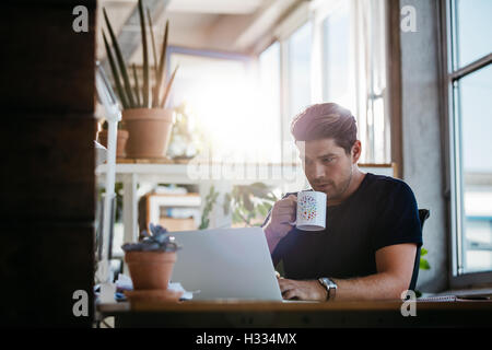 Azienda giovane uomo seduto alla sua scrivania che lavora sul computer portatile e di bere il caffè. Giovane Uomo nel posto di lavoro moderno navigando in internet. Foto Stock