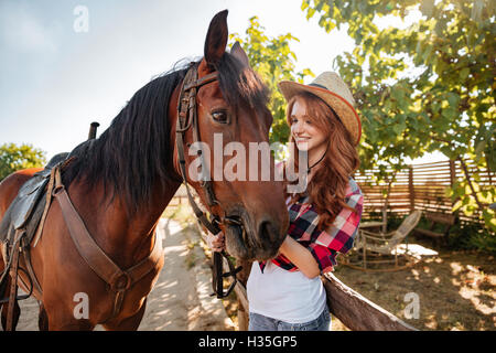 Felice bella giovane donna cowgirl in hat sorridente e prendersi cura del suo cavallo Foto Stock