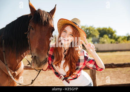 Felice bella giovane donna cowgirl in piedi con il cavallo e la mostra segno di pace Foto Stock