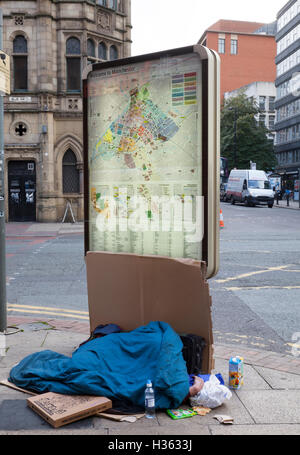 People & i passanti a piedi passato senzatetto mendicante, sonno agitato sul marciapiede contro una mappa stradale affissioni per le strade di Manchester, UK. Foto Stock