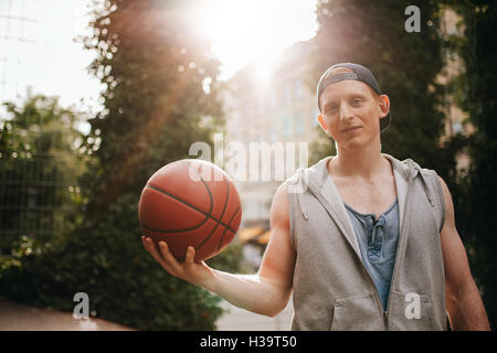 Ritratto di bel giovane azienda una palla da basket sulla corte esterna. Teenage streetball player guardando la fotocamera con una sfera in Foto Stock