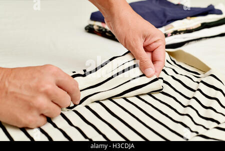 Primo piano di un giovane uomo caucasico il ripiegamento di un polo shirt con un mucchio di vestiti piegati in background Foto Stock