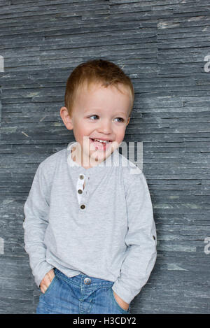 Little Boy appoggiata contro la parete con le mani nelle tasche, sorridente allegramente Foto Stock