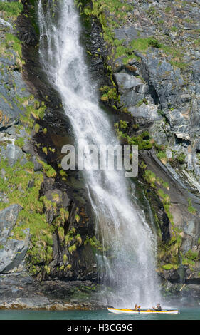 Due persone pausa nell'imbottitura per ammirare una cascata a cascata verso il basso un verticale scogliera in Prince William Sound, Alaska. Foto Stock