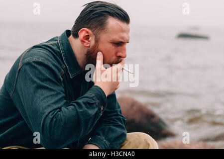 Grave uomo barbuto di fumare una sigaretta sulla spiaggia Foto Stock