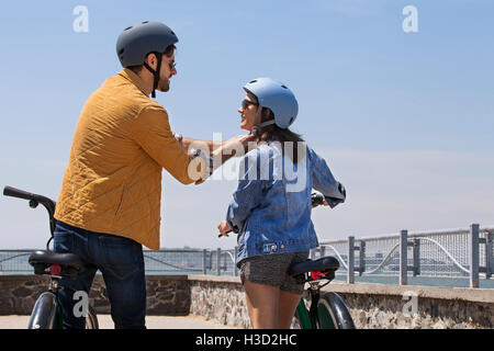 Vista posteriore dell'uomo aiutare la donna a indossare il casco in bicicletta sul lungomare Foto Stock