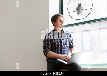 Imprenditore pensieroso seduto con il portatile sul davanzale di office Foto Stock