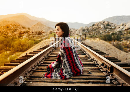 Considerato giovane donna avvolta in una coperta seduti su binari ferroviari con le montagne sullo sfondo Foto Stock