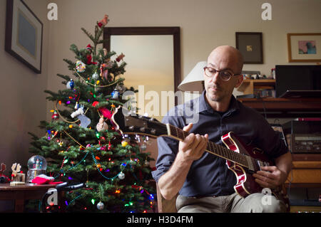 Uomo che suona la chitarra seduto accanto all'albero di natale a casa Foto Stock