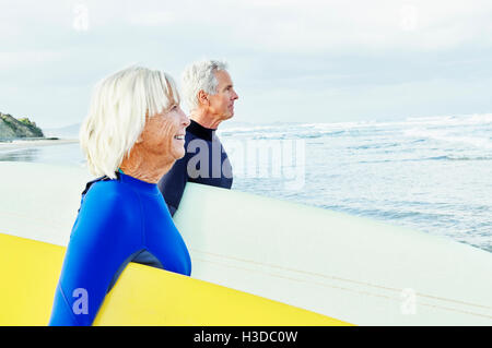 Senior senior donna uomo in piedi su una spiaggia, indossa una muta e il trasporto di tavole da surf. Foto Stock