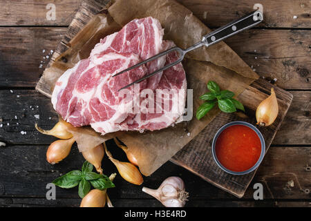 Materie bistecca di carne di maiale sul tagliere con scalogno cipolla, salsa di pomodoro per la marinata, basilico e condimento, servita con carni forcella Foto Stock