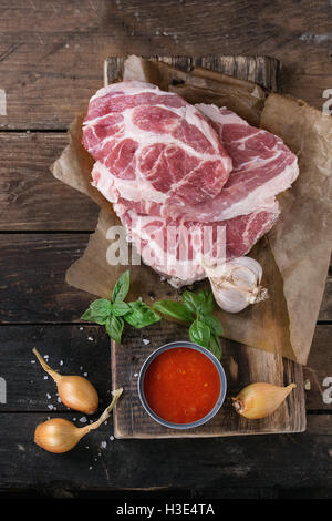 Materie bistecca di carne di maiale sul tagliere con scalogno cipolla, salsa di pomodoro per la marinata, basilico e condimento, servita con carni forcella Foto Stock