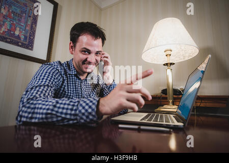 Imprenditore parlando al telefono cellulare, puntare il dito a voi, sorriso seduto alla scrivania in ufficio , utilizzando il telefono cellulare, bel giovane uomo d affari di effettuazione della chiamata, guardando alla fotocamera Foto Stock