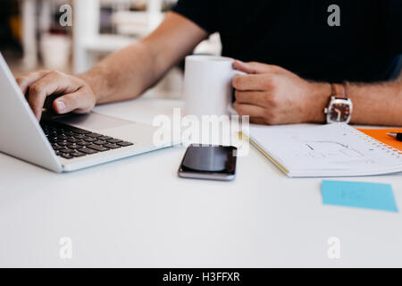 Immagine ritagliata del giovane uomo che lavora sul computer portatile con caffè in mano. Uomo d'affari alla scrivania con il diario e il telefono cellulare. Foto Stock