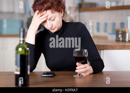 Addolorato giovane donna di bere il vino rosso e la lettura di cattive notizie sul suo telefono cellulare Foto Stock