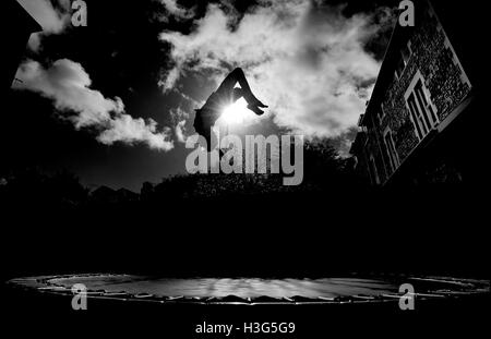 Ragazzo in silhouette, capovolto mentre trampolining in un giardino urbano contro un sole luminoso. *Può essere modello rilasciato su richiesta di Foto Stock