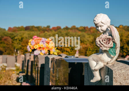 Statua di un angelo tenendo una rosa seduto su una lapide in un cimitero con fiori e lapidi in background Foto Stock