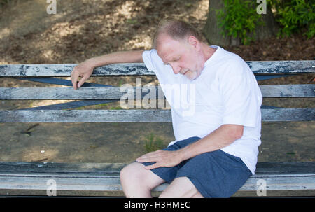Closeup portrait bald old man in white shirt, blue shorts, exhausted, resting on a bench, looking down Foto Stock