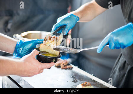 Cheeseburger con carne di maiale tirato essendo posto su burger bun con cetriolini in nero di un contenitore in plastica per alimenti detenute da mani con guanti Foto Stock