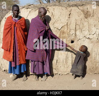 Masai famiglia in abiti tradizionali con un bambino piccolo tirando a madre di vestiti per ottenere attenzione,in piedi di fronte a capanna di fango. Foto Stock