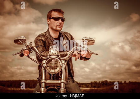 Biker uomo che indossa una giacca di pelle e occhiali da sole seduto sulla sua moto. Filtro applicato in post-produzione. Foto Stock