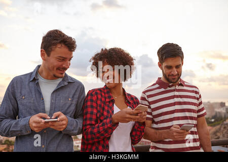 Tre cool giovani amici di ridere mentre guardando i loro telefoni cellulari con uno sfondo bianco, indossando un abbigliamento casual Foto Stock