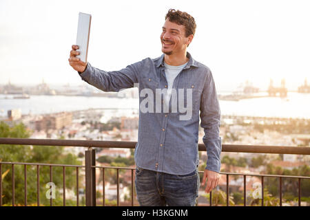 Felice giovane uomo su un ponte che sovrasta la città, prendendo un selfie sul suo tablet indossando un abbigliamento casual Foto Stock