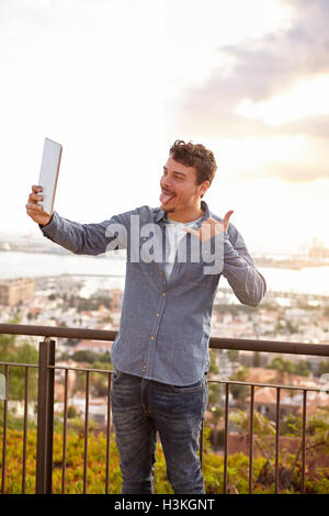 Giovane uomo tirando i volti al suo tablet e indicando un gesto di telefono mentre sta in piedi su un ponte Foto Stock