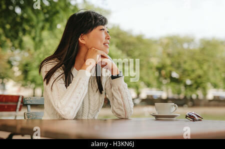 Felice giovane donna seduta al caffè con una tazza di caffè sul tavolo e guardando lontano. Femmina cinese di trascorrere il tempo libero a c esterna Foto Stock