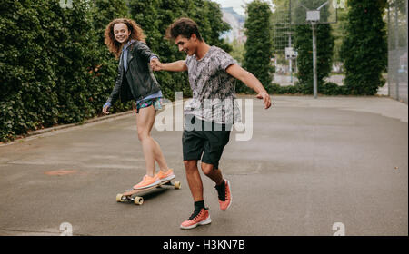 Giovane donna in piedi su un longboard con il supporto dal suo fidanzato. Donna pattinaggio su un campo da basket con un amico. Foto Stock