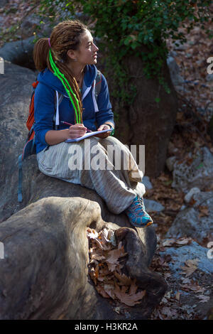 Carino ragazza seduta nella foresta e schizzo di disegno nella sua nota prenota Foto Stock