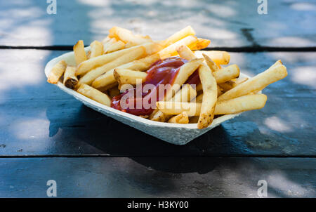 Freschi patate fritte con ketchup nel contenitore di carta sulla tavola di legno in parziale di luce e ombra all'aperto, visto dal lato. Foto Stock