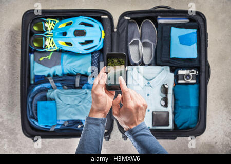 Vista aerea delle mani dell'uomo utilizza lo smartphone al di sopra di valigia con blue bicicletta, casco, zaino, fotocamera retrò e camicia Foto Stock